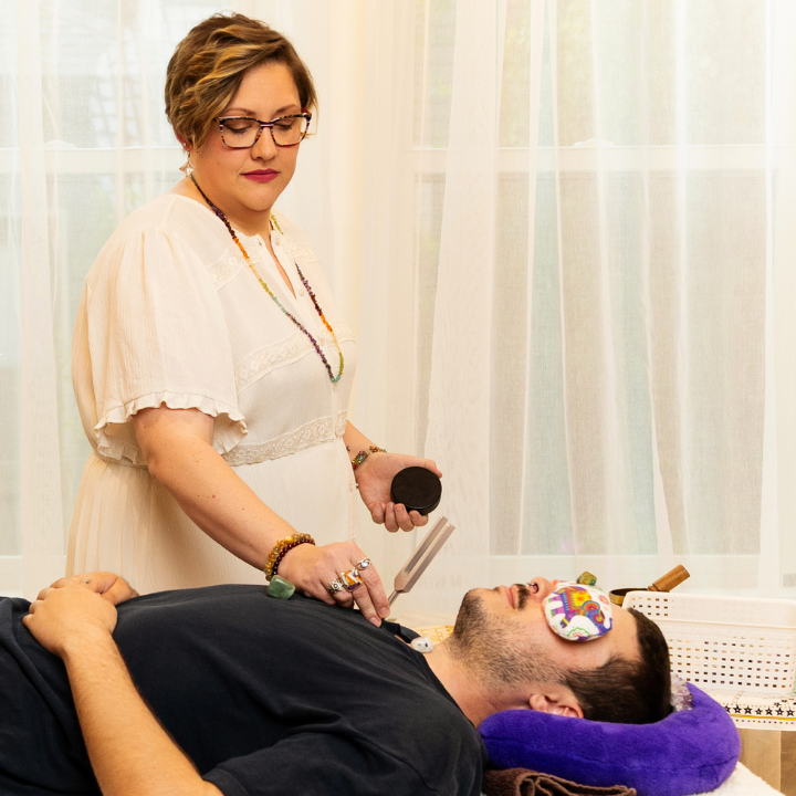 A woman in a white blouse stands beside a reclining man on a massage table, holding a small device over his chest. The man has an eye patch over one eye.